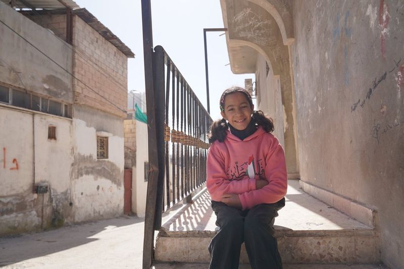 Still from Women in Exile: a girl in a pink hoodie smiling on a doorstep in the camp